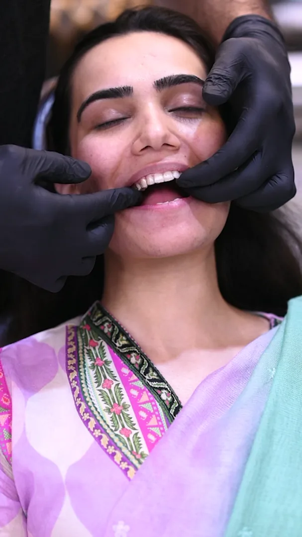 Smiling woman in a dental chair as black-gloved hands hold her lips back to show her upper teeth